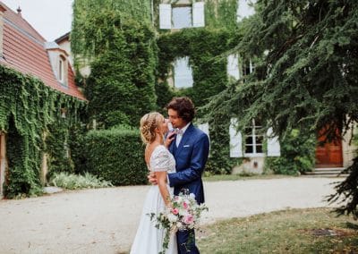 Séance photo des mariés dans le parc du Château de Césarges, avec la façade en fond. Le marié a un costume bleu électrique trois pièces et la mariée une robe bohème et un bouquet de fleurs rose et blanc.