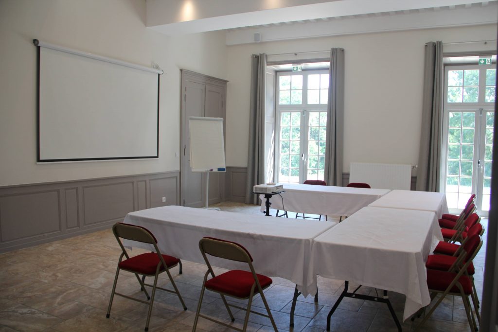 Salle de réunion composé d'un écran de vidéo projecteur, d'un paper board. Les tables sont disposées en U avec des chaises rouges.